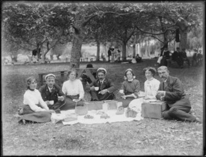 Unidentified group picnicking in a park, showing young men and women sitting around food which has been laid out on a cloth, probably Christchurch district
