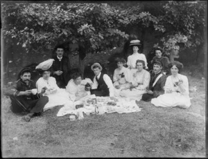Group picnicking, showing unidentified men, some in military uniforms, and women in tea dresses, sitting around a cloth laid with food, possibly Christchurch district