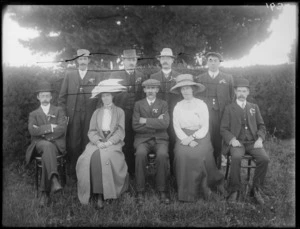 Group of unidentified men and women, in an outdoor location, one women wears a large picture hat and a duster coat, and the men each wear a cluster of ribbons pinned to their lapel, probably Christchurch district