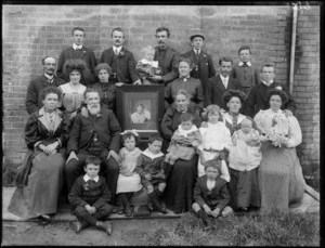 Family group [reunion?], showing unidentified men, women and children with framed photograph of an absent member [memento mori?], possibly Christchurch district