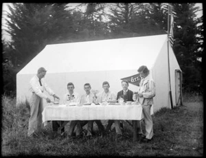 Unidentified young men in formal wear, sitting at table ready to have a meal, next to tent 'Lily' at camp site, probably in Sumner, Christchurch