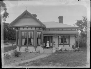 Exterior view of single storied house, shows unidentified man and young girl on the porch with a pram on the grass, probably Christchurch district