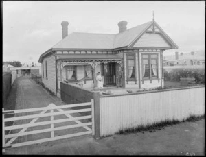 Exterior view of a single storied house, shows unidentified couple on porch and a gated driveway, probably Christchurch district