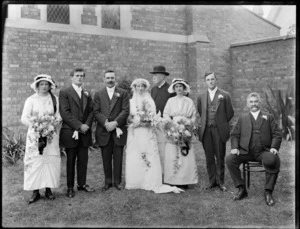 Wedding party portrait outside a church, unidentified bride and groom with family members and an older priest with bowler hat, women holding flowers, probably Christchurch region