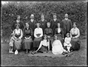 Unidentified group portrait of men, women and children on grass in front of a hedge, probably Christchurch region