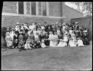 Large wedding group portrait outside a church, unidentified bride and groom with extended family, an older priest, children and babies in front, probably Christchurch region