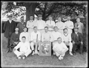 Portrait of unidentified men's cricket team and coaches, players in uniform, one with cricket bat, under a tree with a large shield trophy wall plaque, [Hagley Park?], Christchurch