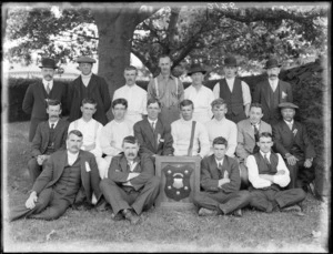 Portrait of unidentified men's cricket team, some with ribbons, one with cricket bat, under a tree with a large shield trophy wall plaque, [Hagley Park?], Christchurch