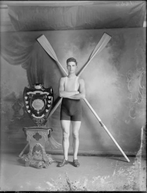 Studio portrait of unidentified male rower, winner of The Cleary competition Shield, including shield and oars crossed diagonally behind, probably Christchurch district