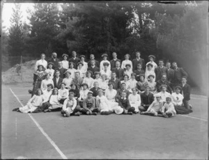 Group portrait of unidentified youngish men and women with children, on a tennis court in front of trees, probably Christchurch region