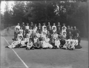 Group portrait of unidentified youngish men and women with children, on a tennis court in front of trees, probably Christchurch region