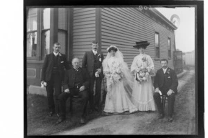 Wedding party portrait by the corner of a house, unidentified bride with long veil and groom with family members, women holding flowers, probably Christchurch region