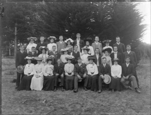 Group portrait of unidentified youngish men and women, some with carnations [wedding group?], in front of trees, women wearing hats, probably Christchurch region