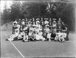 Group portrait of unidentified youngish men and women with children, some with carnations [wedding group?], women wearing hats, on a tennis court in front of trees, probably Christchurch region