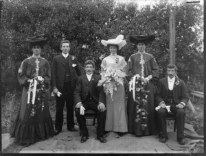 Wedding party portrait in front of hedge, unidentified bride and groom with family members, women with flowers and fine hats, probably Christchurch region