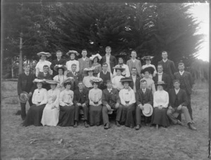 Group portrait of unidentified youngish men and women, some with carnations [wedding group?], in front of trees, women wearing hats, probably Christchurch region