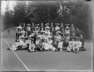 Group portrait of unidentified youngish men and women with children on a tennis court in front of trees, women wearing hats, probably Christchurch region