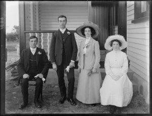 Wedding party portrait in front of the entrance of a house, unidentified bride and groom standing with best man and bridesmaid sitting, probably Christchurch region