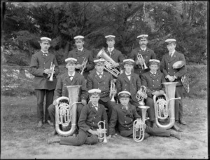 Group portrait of unidentified brass band members in uniform and hats with insignia, holding their instruments, on a lawn in front of trees, probably Christchurch region
