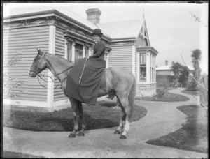 Unidentified woman in riding attire, with bowler hat and riding crop, sitting side saddle on her horse, in front garden with wooden house behind, probably Christchurch region