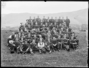Large group portrait of unidentified men on a hillside with cricket bats and musical instruments, wearing ribbons or medals, with hilly farmland beyond, probably Christchurch region