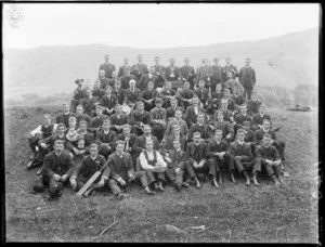 Large group portrait of unidentified men on a hillside with cricket bats and musical instruments, wearing ribbons or medals, with hilly farmland beyond, probably Christchurch region