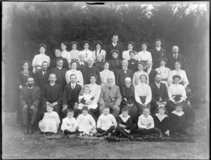 Large group portrait of unidentified adults, with children sitting in front, with an elderly couple in the centre, hedge beyond, probably Christchurch region