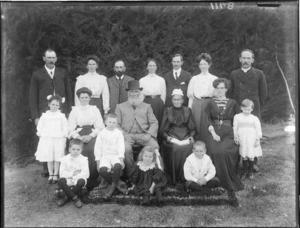 Family portrait, unidentified grandparents with their adult family members and grandchildren, with hedge beyond, probably Christchurch region