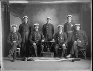 Studio portrait of the unidentified Christchurch Fire Brigade members in dress uniform and caps, some with lapel medals, with stretcher and hamper in front