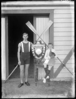 Unidentified single coxed scull rowing team, rower holding a shield trophy with young boy coxswain, crossed oars behind, outside the Avon Rowing Club, Christchurch