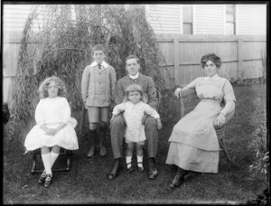 Family portrait of unidentified young couple sitting in cane chairs in the backyard with their three children, probably Christchurch region