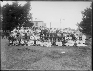 Large group portrait of men, women and children, in paddock in front of house and windmill, probably Christchurch district