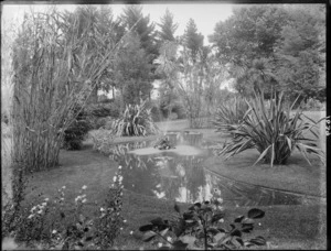 Park or garden, with a pond and fountain, surrounded by flax bushes, bamboo and lawn, with trees beyond, probably Christchurch region