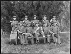 Group portrait of unidentified brass band members in military style uniforms and slouch hats, holding their instruments, on a lawn in front of trees, probably Christchurch region