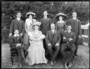 Family portrait, unidentified older couple with family members, four men, two women and a boy [attending a wedding?], women with hats and boa feather scarves, trees beyond, probably Christchurch region