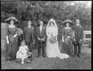 Wedding party portrait, unidentified bride in long veil and groom with bridesmaids and groomsmen and young flower girl, pine trees beyond, probably Christchurch region