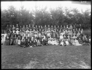 Very large wedding group portrait in front of pine trees, unidentified bride and groom with extended family with children in front, women wearing hats, probably Christchurch region