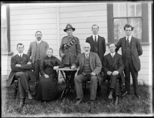 Family group portrait, unidentified elderly couple with six men, the youngest man in military uniform with swagger stick and bandolier over left shoulder, wooden building behind, probably Christchurch region