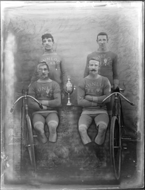 Studio portrait, unidentified Dunedin four man Dunlop Cycling Team, with cup and two racing bikes, probably Christchurch region