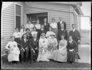 Wedding group portrait by the rear entrance of a wooden house with budgie cage, unidentified bride and groom with extended family, including young girl and baby, probably Christchurch region
