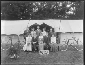Group of unidentified young men and women in front of two tents with overlapping awning, with bicycles and 'Imperial Camp' sign, trees beyond, [Sumner?], Christchurch