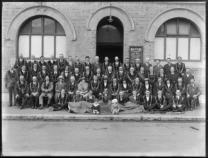 Group portrait of unidentified Hibernian Australasian Catholics Benefit Society members, in front of Saint Patrick's Branch No 82 hall, young boy and girl in front sitting on large flag with emblem, probably Christchurch