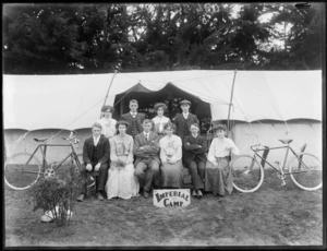 Group of unidentified young men and women in front of two tents with overlapping awning, with bicycles and 'Imperial Camp' sign, trees beyond, [Sumner?], Christchurch