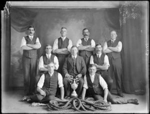 Studio portrait of a [tug-of-war?] team, showing eight unidentified members and coach, with coiled hawser laid rope in front and cup, Christchurch