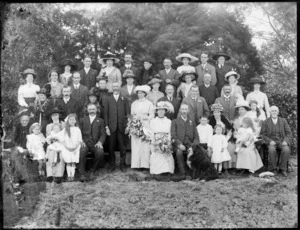 Large wedding group portrait on rough ground in front of trees, unidentified bride and groom with extended family, children and a dog in front, women with hats, probably Christchurch region
