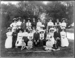 Group portrait of unidentified adults with hats and children with white caps, on grass with trees and river behind, with cricket bat and rowing oars in front, probably Christchurch region