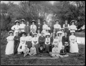 Group portrait of unidentified adults with hats and children with white caps, on grass with trees and river behind, with cricket bat and rowing oars in front, probably Christchurch region
