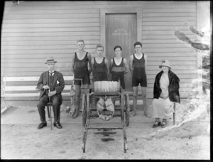 Group of unidentified young male lifesavers in bathing suits, includes elderly couple and a life saving reel, with writing on a door saying 'Private Members Only', probably Christchurch district