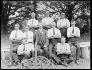 Group of unidentified men outdoors, probably Christchurch district, showing man seated in front holding an unidentified object, with coiled rope by his feet