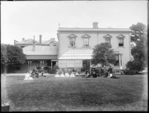 Unidentified group of adults and children sitting on a large lawn beside a large wooden building with a glass conservatory, probably Christchurch district